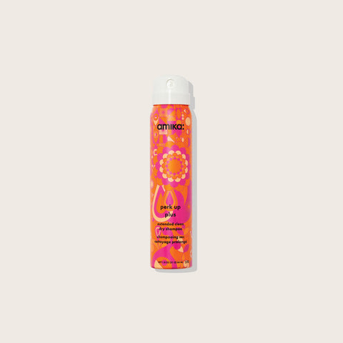 Flacon de shampooing sec Amika Perk Up Plus pour cheveux, bouteille blanche avec étiquette colorée, à côté d'un fond neutre.